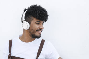 Glücklicher junger Mann mit Kopfhörern, der vor einer Wand Musik hört - JCZF01153