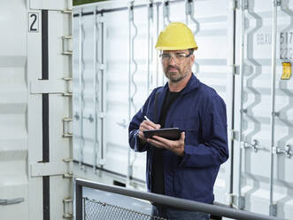 Foreman looking at cargo container writing with digitzed pen on digital tablet - CVF02232