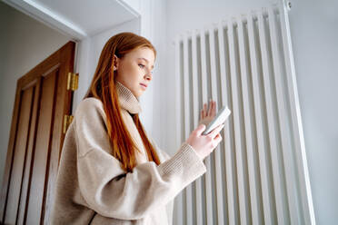 Teenager-Mädchen, das mit einem Mobiltelefon die Temperatur des Heizkörpers zu Hause überprüft - MDOF00537