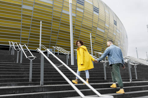 Lächelnde Frau hält die Hand eines Mannes und geht auf den Stufen vor dem Stadion - VIVF00320