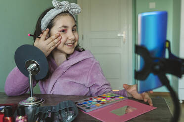 Smiling girl filming make-up vlog through smart phone at home - OSF01364
