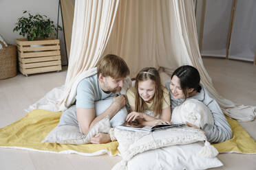 Lächelnde Eltern und Tochter beim Lesen eines Buches in einem Deckenzelt - EYAF02532