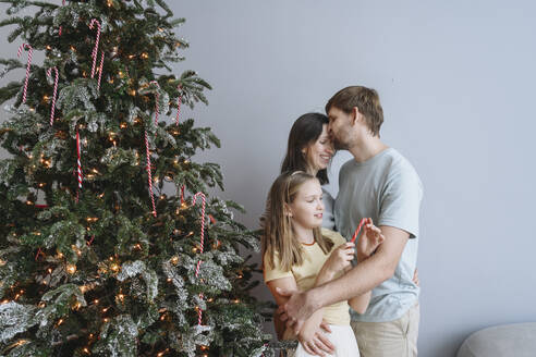 Man kissing woman on forehead with daughter by Christmas tree at home - EYAF02552