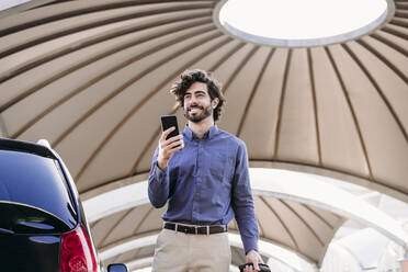 Happy young businessman with smart phone standing at parking lot - EBBF07746