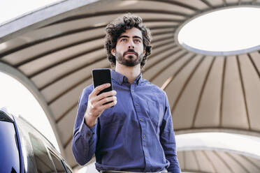 Young businessman with smart phone standing under ceiling in parking lot - EBBF07747