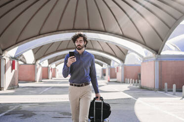 Junger Geschäftsmann geht mit Gepäck und benutzt ein Smartphone - EBBF07748