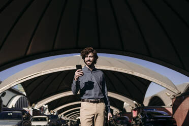 Smiling businessman with smart phone standing in parking lot - EBBF07751