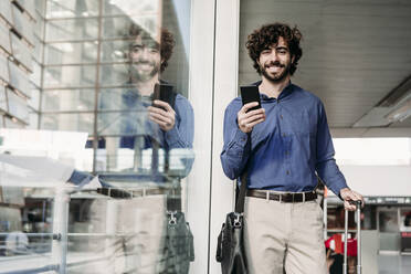 Happy businessman standing with smart phone at railroad station - EBBF07757