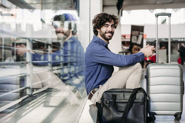 Lächelnder junger Geschäftsmann mit Gepäck und Smartphone auf dem Bahnhof - EBBF07760