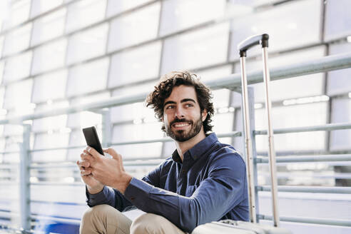 Lächelnder junger Geschäftsmann sitzt mit Smartphone am Bahnhof - EBBF07763