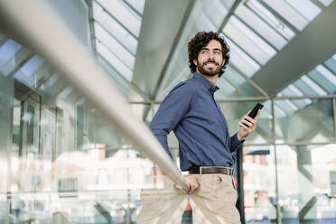 Contemplative businessman with smart phone standing by railing - EBBF07776