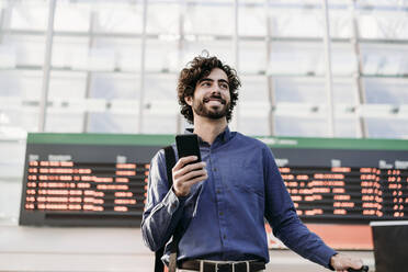 Happy businessman with smart phone standing in front of arrival departure board - EBBF07784