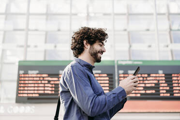 Happy businessman using mobile phone by arrival departure board at station - EBBF07786