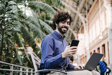 Happy businessman with laptop using smart phone sitting on bench - EBBF07791