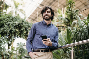 Smiling businessman with smart phone standing by railing in front of plants - EBBF07797