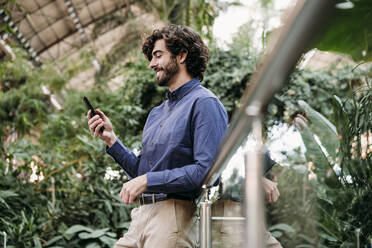 Lächelnder Geschäftsmann, der sein Smartphone am Geländer vor den Pflanzen benutzt - EBBF07798