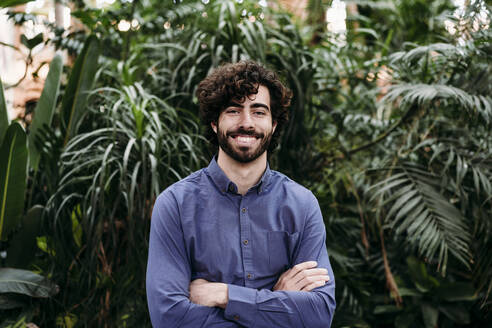 Smiling young businessman standing with arms crossed in front of plants - EBBF07799