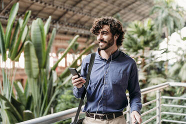 Smiling businessman wearing in-ear headphones standing with smart phone by plants - EBBF07803