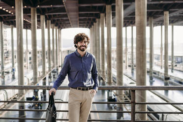 Happy businessman standing with bag in front of railing - EBBF07808