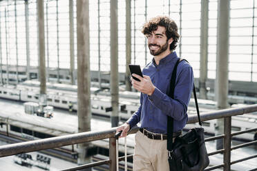 Glücklicher Geschäftsmann mit Smartphone auf einer Brücke am Bahnhof stehend - EBBF07812