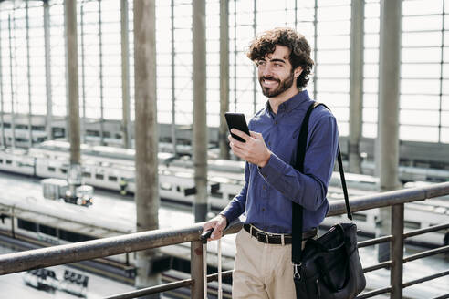 Happy businessman with smart phone standing on bridge at station - EBBF07812