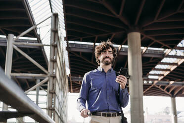 Smiling businessman with smart phone standing at station - EBBF07814