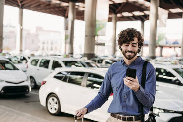 Smiling businessman using smart phone in front of cars - EBBF07819