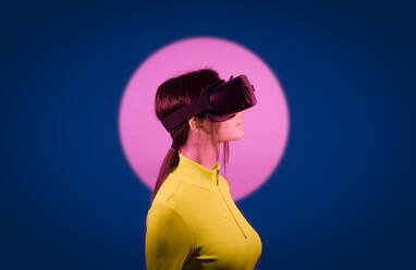 Woman watching on virtual reality goggles by pink light over black background - JSMF02623