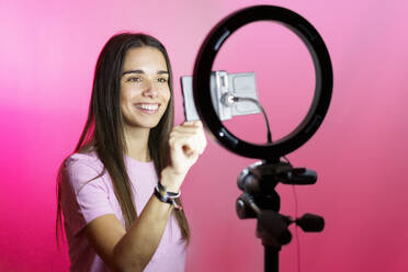 Lächelnder Influencer, der mit seinem Smartphone im Ringlicht vor einem rosa Hintergrund filmt - JSMF02632