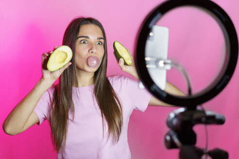 Frau mit Avocadohälften bläst Blase und Vlogging über rosa Hintergrund - JSMF02638