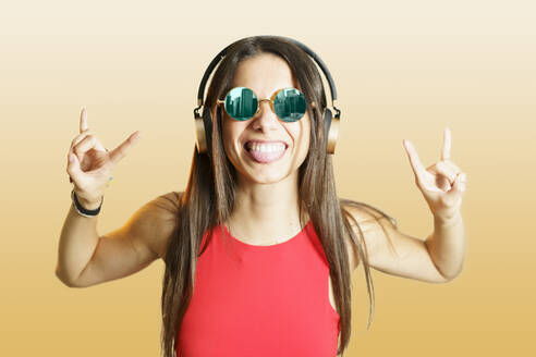 Junge Frau mit Kopfhörern gestikuliert Horn Zeichen über gelben Hintergrund - JSMF02643