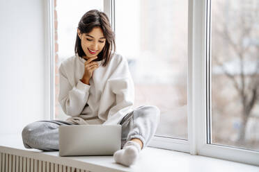 Happy beautiful woman with hand on chin using laptop in front of window - MDOF00547