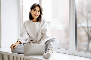 Happy woman sitting with laptop on window sill - MDOF00548