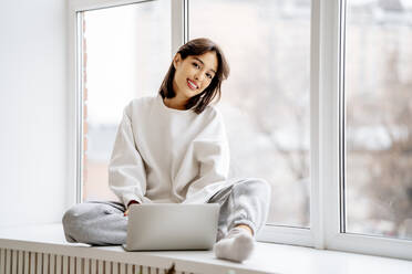 Happy young woman sitting with laptop on window sill - MDOF00549