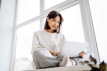 Smiling young woman sitting with laptop in front of window at home - MDOF00551