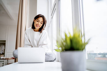 Lächelnde junge Frau mit Laptop auf der Fensterbank - MDOF00554