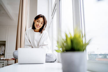 Smiling young woman using laptop on window sill - MDOF00554