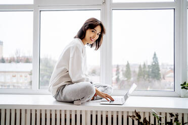 Happy beautiful woman sitting with laptop on window sill at home - MDOF00555