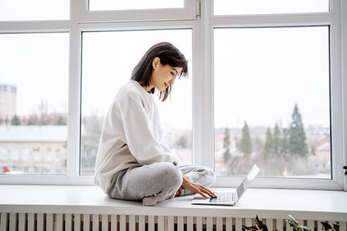 Lächelnde Frau mit Laptop auf der Fensterbank zu Hause - MDOF00556