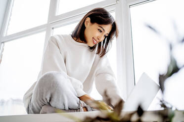 Happy woman using laptop on window sill at home - MDOF00557