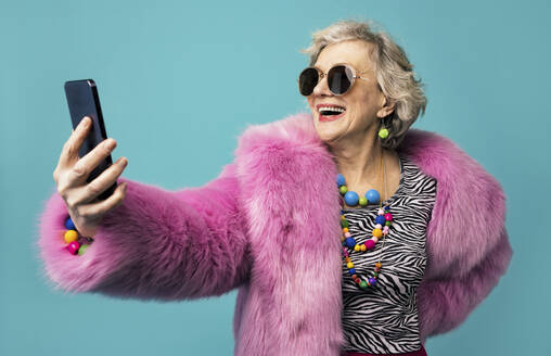 Happy senior woman wearing jewelry taking selfie through smart phone against turquoise background - OIPF02964