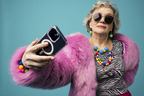 Lächelnde ältere Vloggerin, die ein Selfie mit ihrem Smartphone vor einem türkisfarbenen Hintergrund macht - OIPF02976
