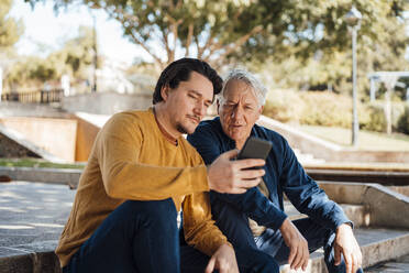 Man showing smart phone to father sitting on steps - JOSEF16271