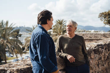 Älterer Mann im Gespräch mit seinem Sohn an einer Mauer stehend an einem sonnigen Tag - JOSEF16282