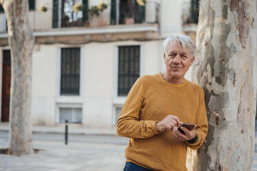 Älterer Mann lehnt mit Smartphone an einem Baum - JOSEF16347