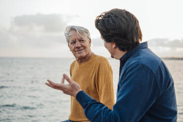 Smiling senior man with son having discussion in front of sea - JOSEF16378