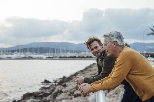 Happy mature man leaning on railing with father in front of sea - JOSEF16394