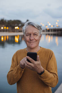 Lächelnder älterer Mann stehend mit Smartphone bei Sonnenuntergang - JOSEF16400