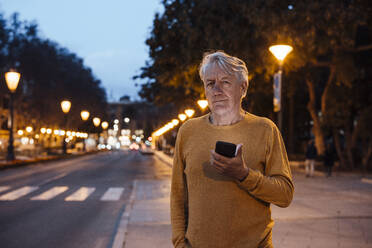 Lächelnder älterer Mann mit Mobiltelefon auf dem Gehweg stehend - JOSEF16404