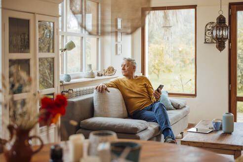 Nachdenklicher älterer Mann sitzt mit Smartphone auf dem Sofa zu Hause - JOSEF16418